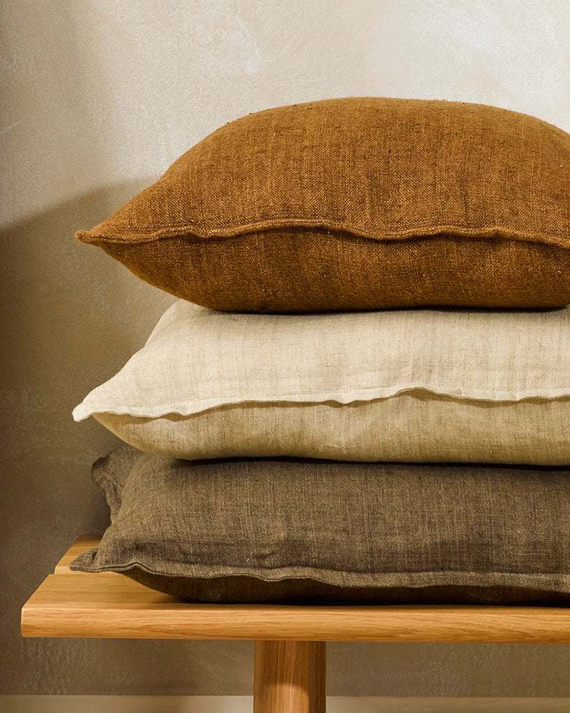 Three Baya linen cushions in brown tones, stacked on a stool