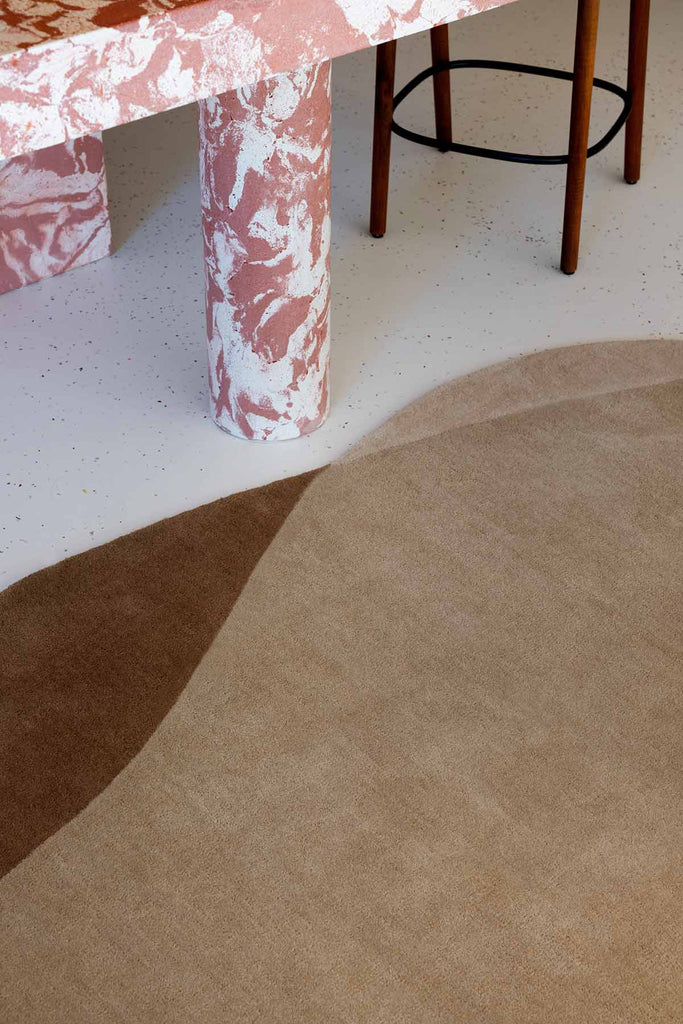 A closer view of a handtufted rug in warm caramel brown tones with an irregular, organic shape. Styled in a modern dining room. By Brink and Campman