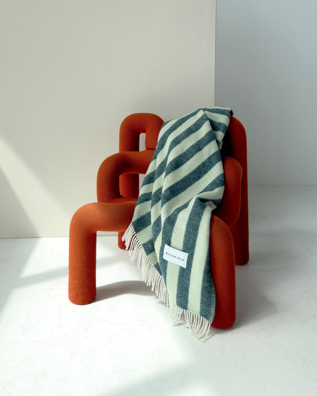 A sage and forest green striped throw blanket by Maison Deux, draped over a modern orange chair. 