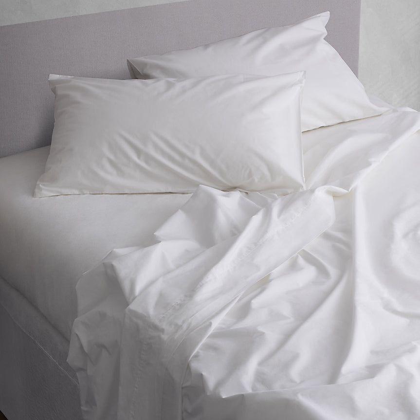 White bedding set in 100% cotton with two pillows and rumpled sheets on a bed against a light gray wall.