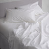 White bedding set in 100% cotton with two pillows and rumpled sheets on a bed against a light gray wall.