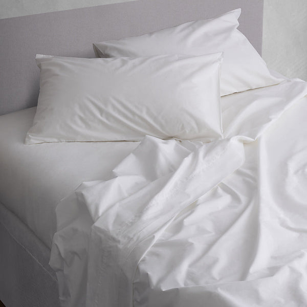White bedding set in 100% cotton with two pillows and rumpled sheets on a bed against a light gray wall.