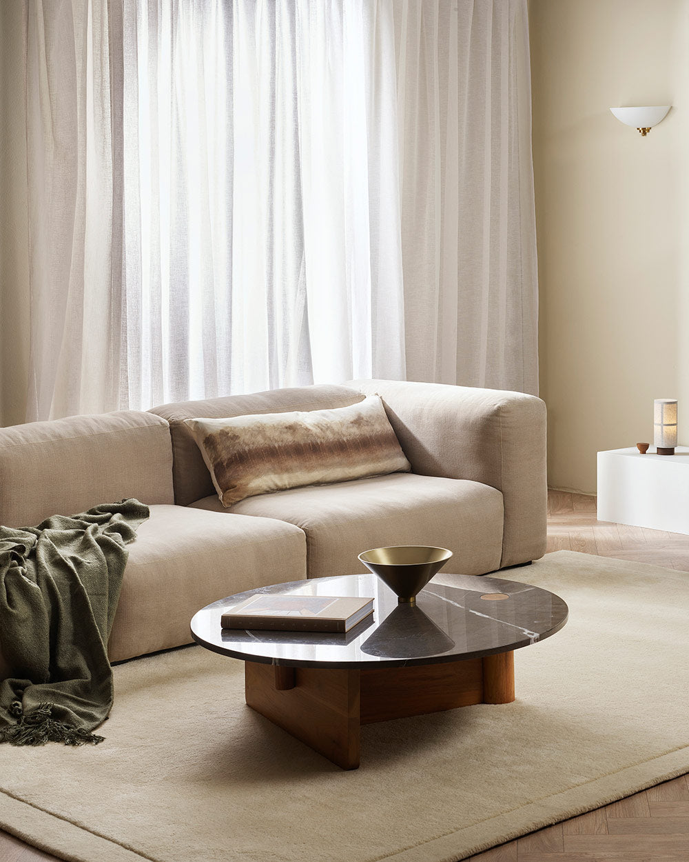 A couch in a stylish modern living room, with a green throw blanket and a gorgeous long lumbar cushion, featuring an abstract forest rendered in soft brown tones. 
