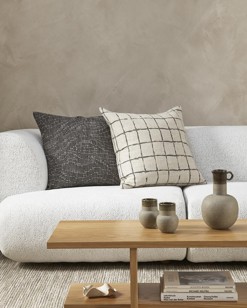 The Baya Harrison cushion in black, featuring a  grid-like design with a handcrafted look on a linen blend base, next to a black textural cushion on a creamy white couch.
