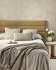 A taupe coloured lumbar cushion featuring a grid design in white in a hand-painted style, the Johnson Taupe is seen here on a bed in a stylish bedroom with other cushions.
