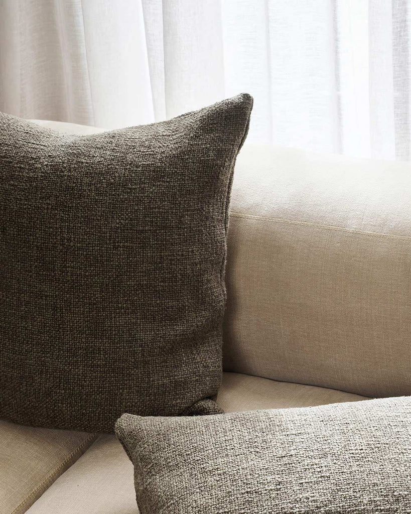 A soft woven textural cushion in tones of deep rosemary green on a cream toned couch, by Baya. 