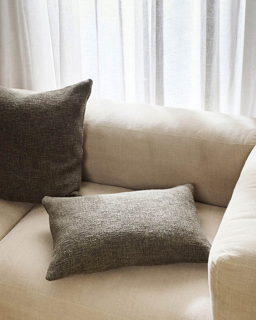 Two textural deep green toned cushions, one large and one lumbar, sitting on a modern style cream toned couch. By Baya. 