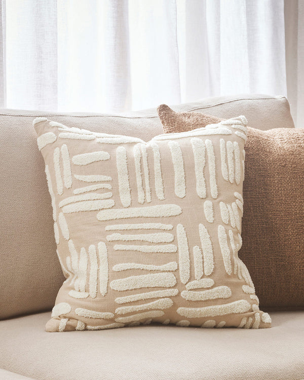 A modern textured cushion with patches of cream embroidery in an irregular striped pattern around the beige body of the cushion, on a cream toned couch. By Baya. 