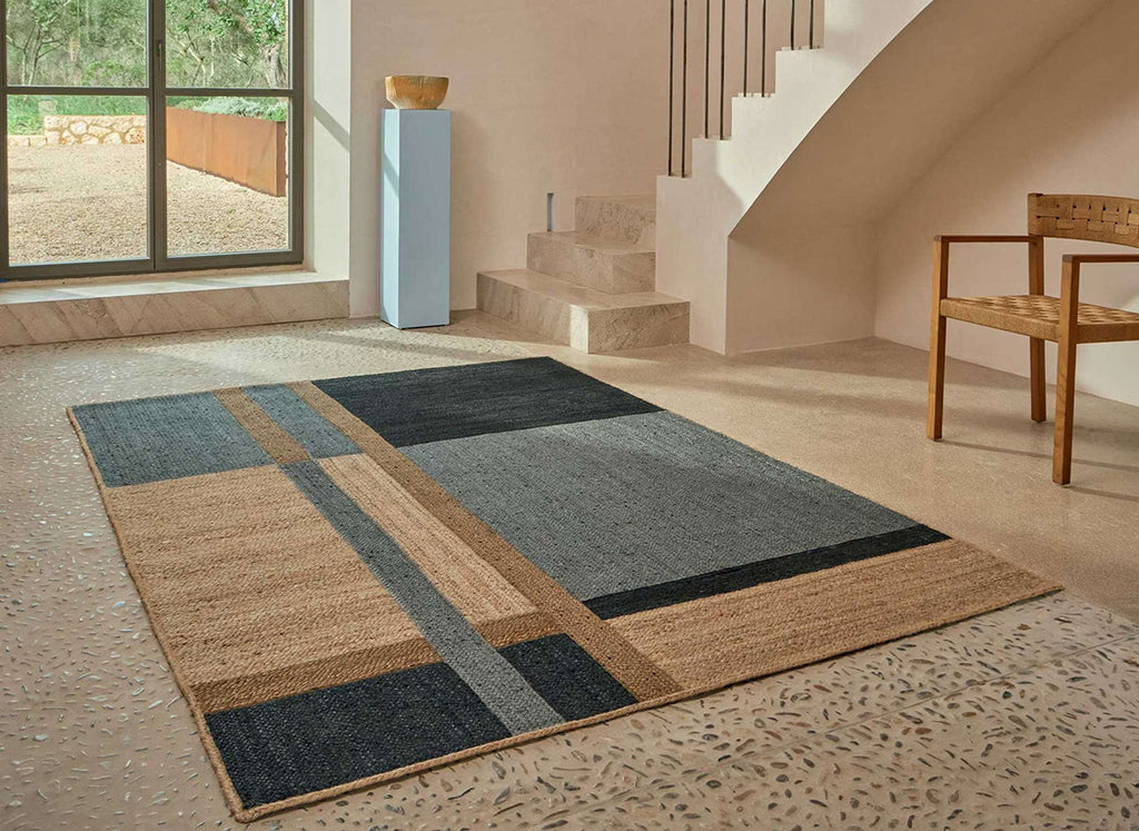 Elitis blue geometric handbraided jute patterned rug on a textured floor with a staircase and window in the background