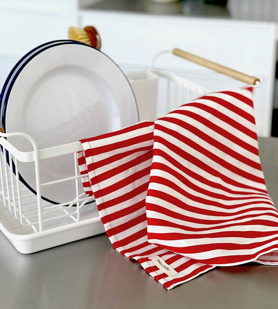 A striped tea towel from Father Rabbit, in the shade French Nautical Red Stripe. It is draped over a dish rack, the red and white stripes add an element of bright fun to the kitchen.