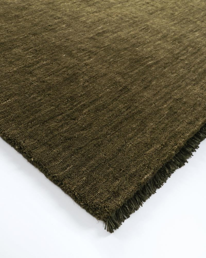 A corner image of a warm deep green toned floor runner with a fringed edge. 