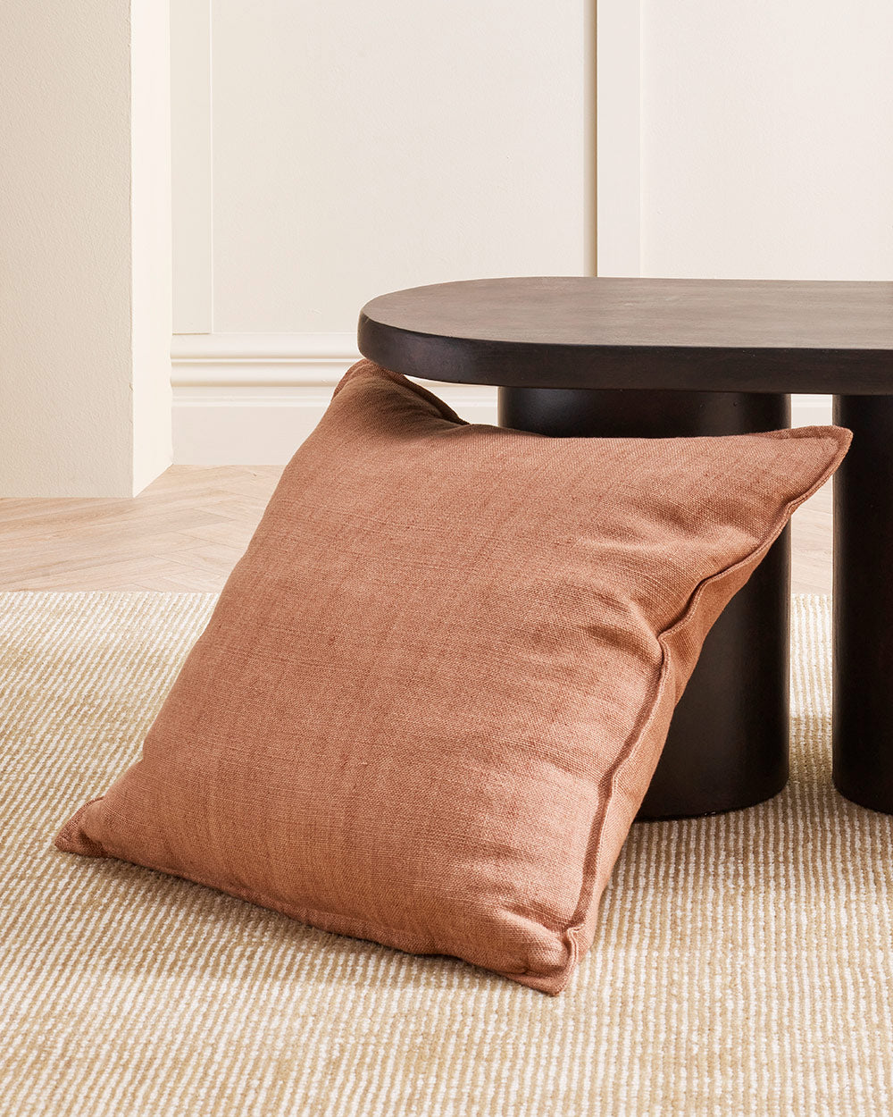 A muted pink linen textured cushion with a small flange edge, leaning against a coffee table. By Baya. 