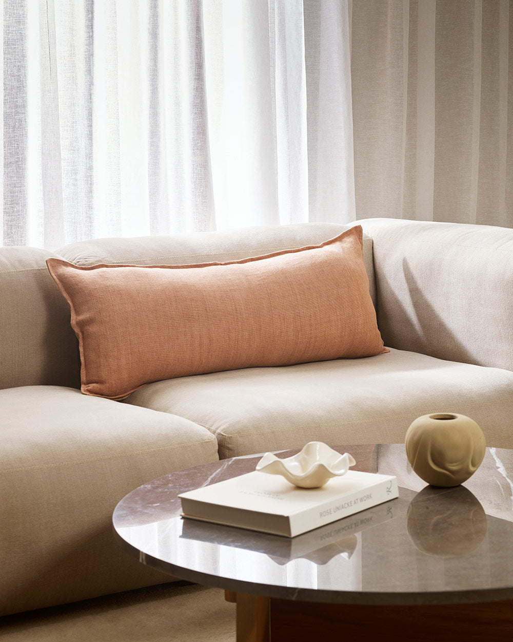 A long lumbar cushion in muted pink on a cream toned couch in a stylish living room by Baya. 