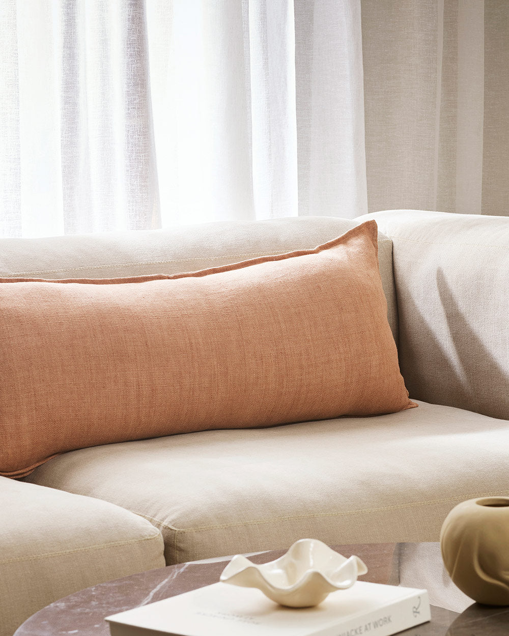 A long lumbar cushion in a dusty clay pink sitting on a cream toned couch. By Baya. 
