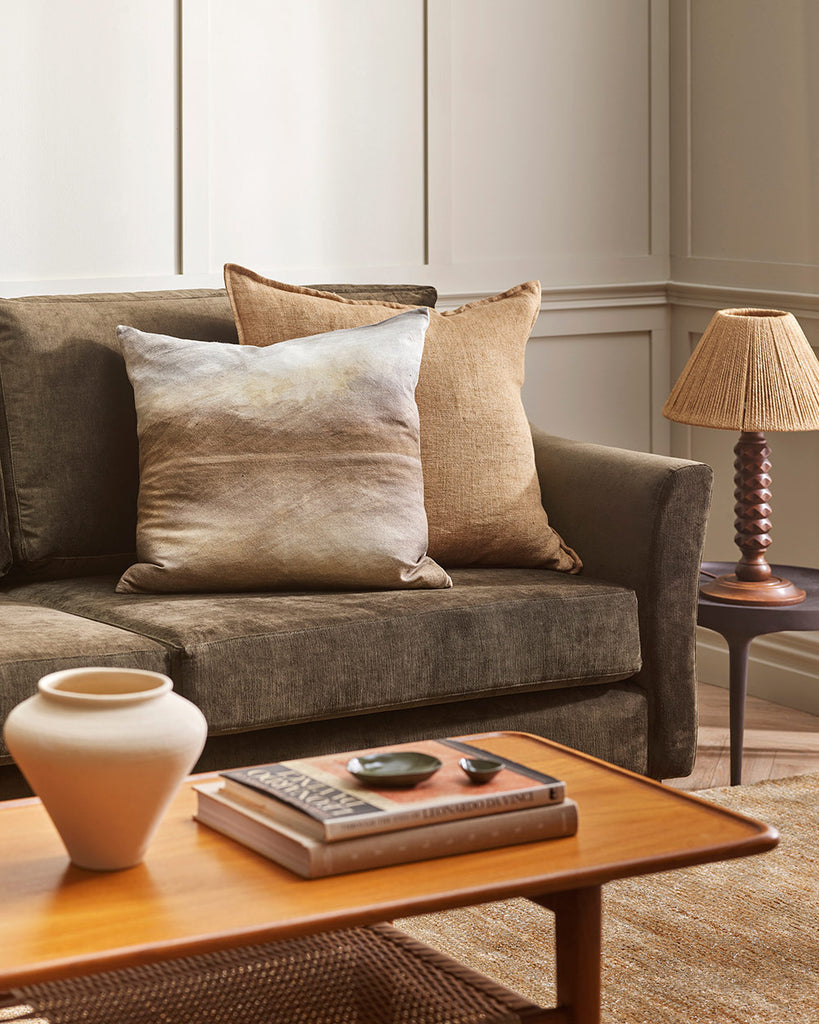 A sandy brown toned cushion next to a handpainted abstract landscape printed cushion, in an earth toned living room by Baya. 