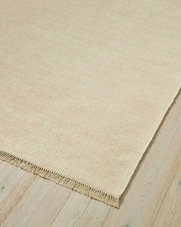 A corner of a 100% linen fibre floor rug in a soft warm cream tone with a matching fringed edge. 