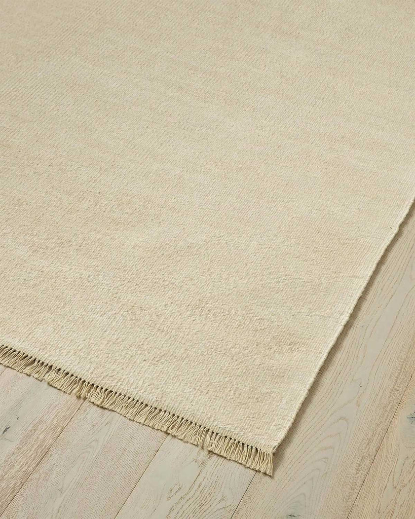 A corner of a 100% linen fibre floor rug in a soft warm cream tone with a matching fringed edge. 