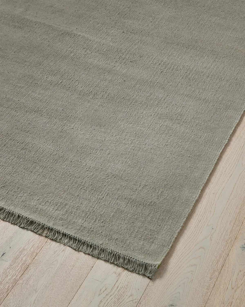 The edge of a blue grey floor rug made from 100% linen fibres with a matching fringed edge. 