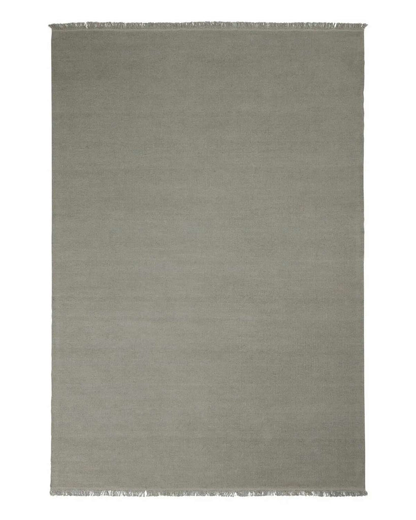 A full image of a handwoven linen floor rug in a soft grey green tone with fringe edging. 