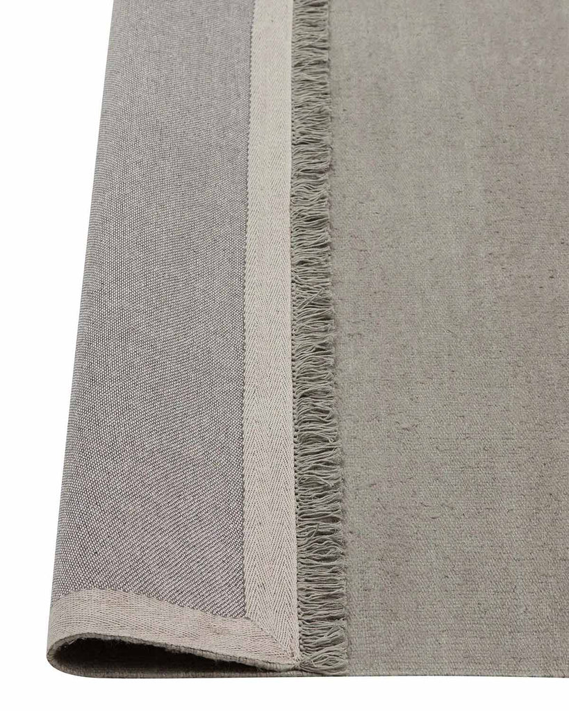 A corner image of a lichen toned grey green floor rug in linen with the edge flipped up to show the underside of the fringed edging and the durable cotton backing. 