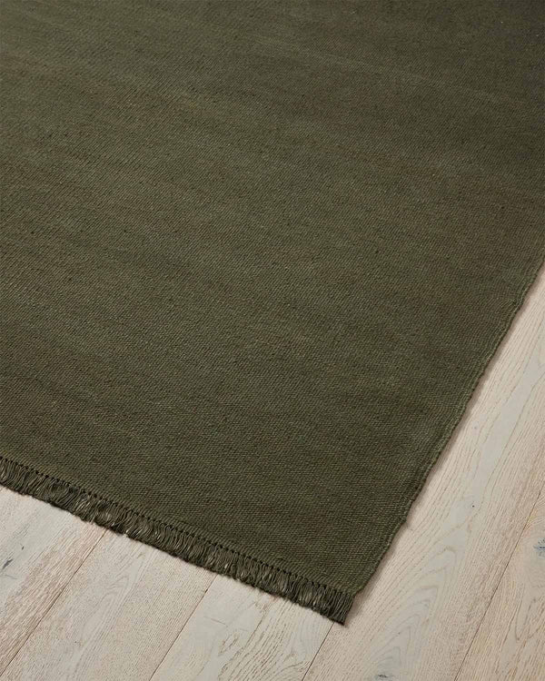 A corner image of a deep olive green floor rug with matching toned fringed edges in 100% linen. 