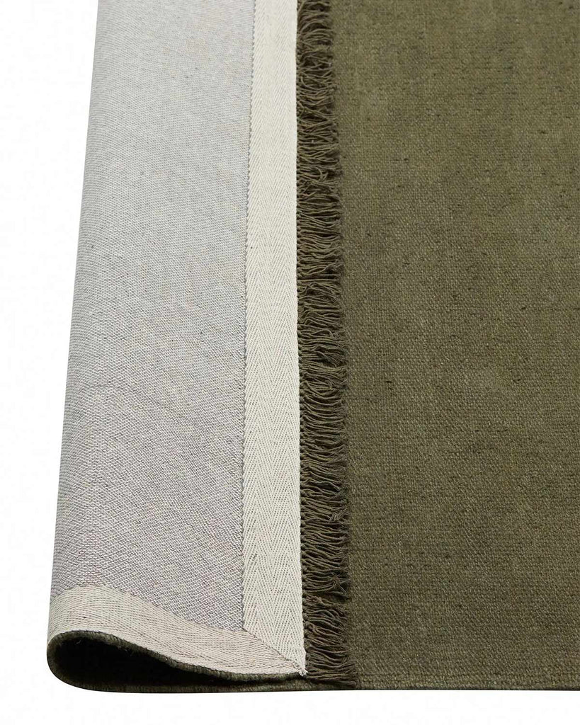 A corner image of a dark green floor rug in 100% linen, with the edge flipped up to show the durable cotton backing and the underside of the fringe edging. 