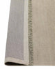 A handmade linen floor rug in cool taupe with fringed edges— the corner is flipped up to show the durable cotton underside. 
