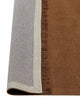 A corner image of a warm brown floor rug made with linen fibres with the edge flipped up to show the underside of the fringe and the durable cotton backing. 