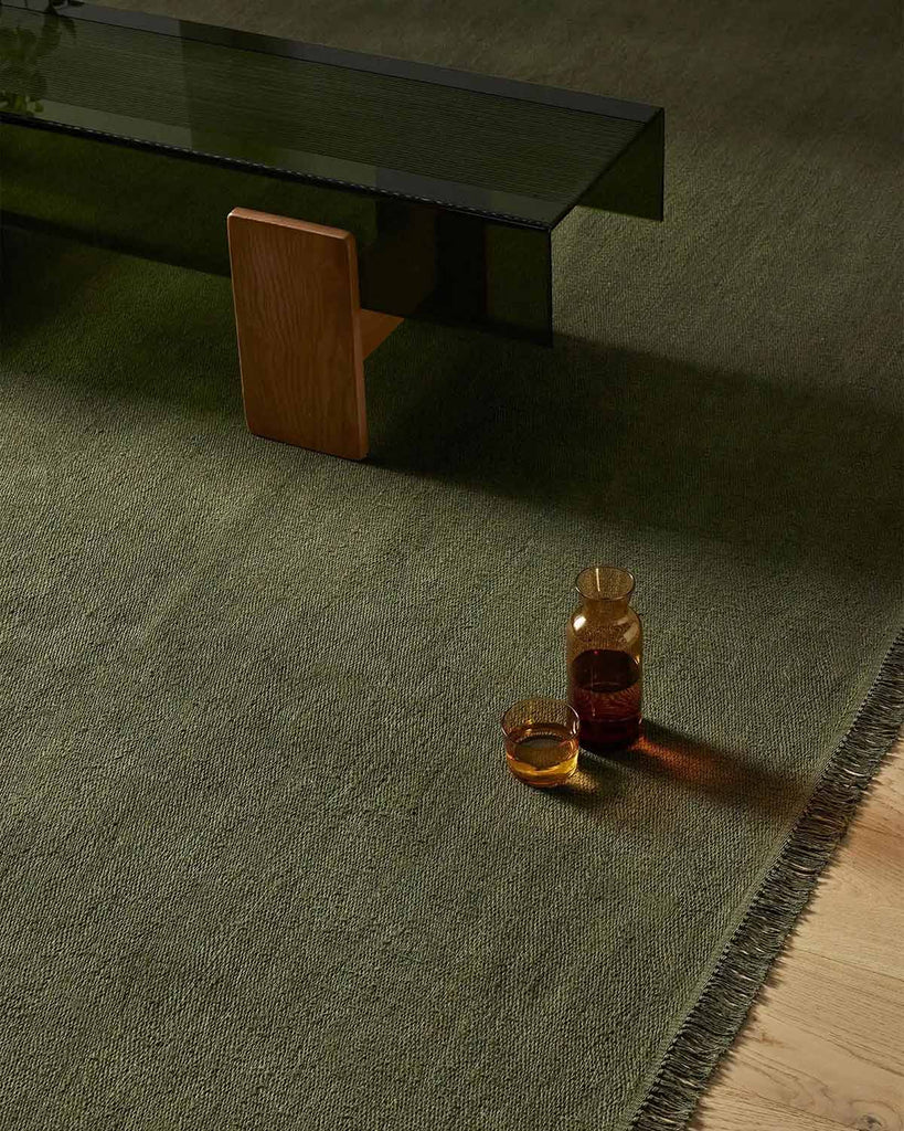 A large moss green linen fibre floor rug with fringe edging and a carafe made of smoked glass. 