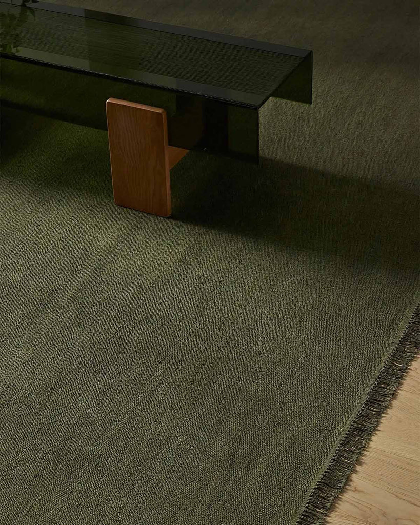 A handwoven linen fibre floor rug in olive green with fringed edges, with a modern style coffee table sitting on it. 