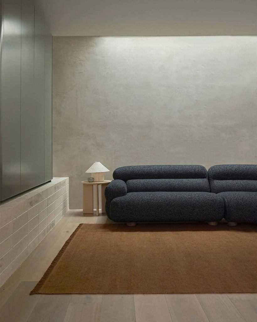 A modern style sitting area with a grey couch and a warm brown floor rug made with linen with fringed edges. 