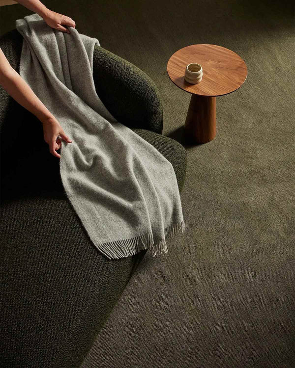 A modern sitting area with a coffee table and armchair atop a large olive green linen floor rug. 