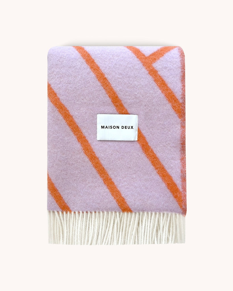 A folded, line patterned throw blanket in shades of lilac and vibrant orange, by Maison Deux