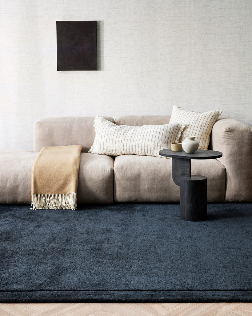 A cool toned lounge with an ink blue floor rug, a white couch and a warm sandy beige throw blanket. 