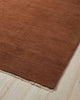 A corner image of a rust orange brown floor rug handwoven from 100% NZ wool, with cream fringed edging. 