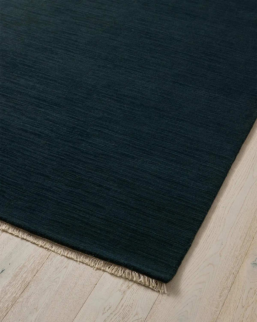 A corner of a deep ink blue floor rug in 100% wool with cream fringed edging. 