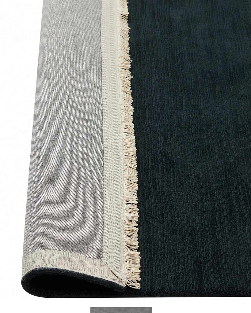 A deep blue 100% wool floor rug with off-white fringed edges, the corner of which is flipped up to show the cotton backing. 