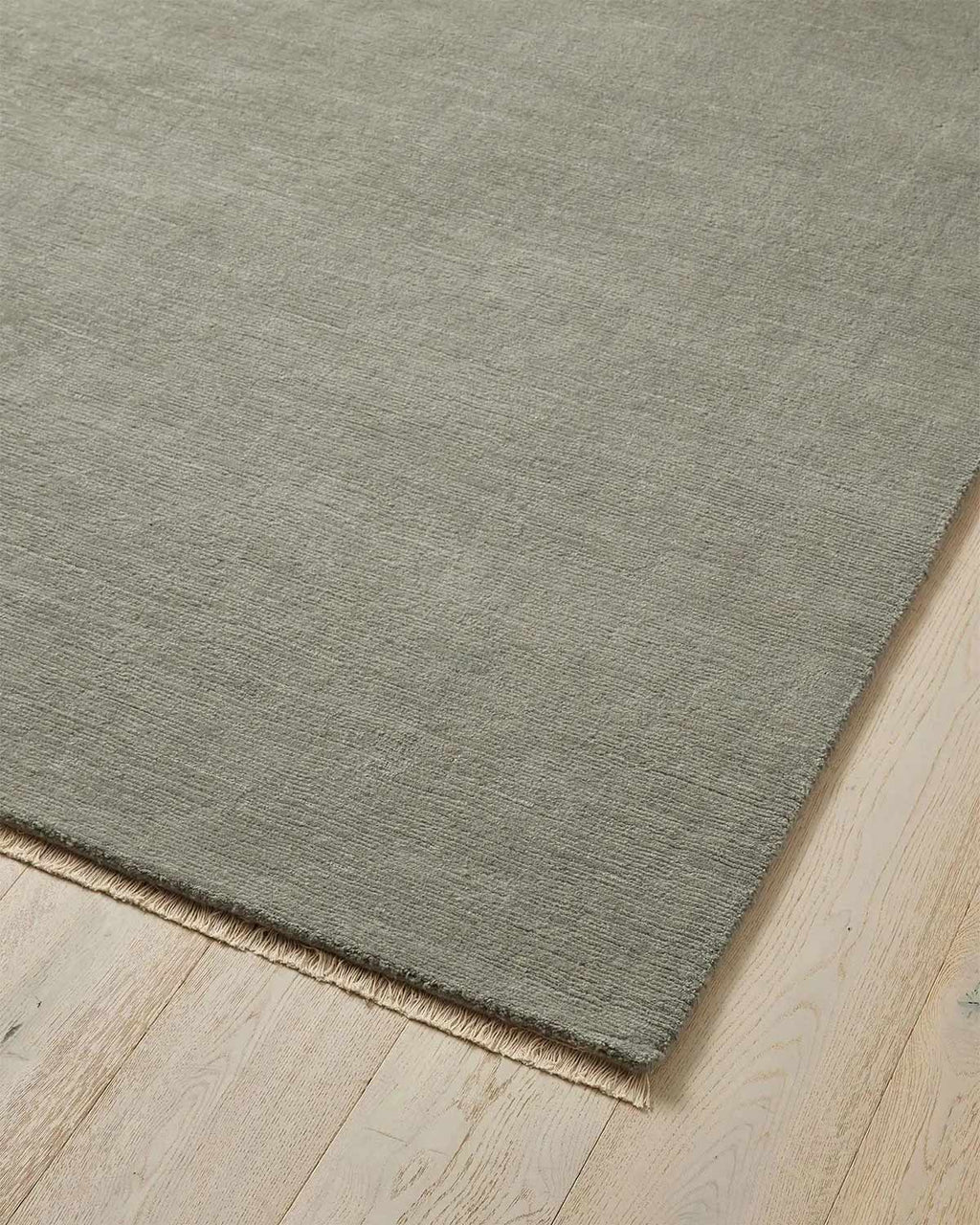The corner of a grey green floor rug with a linear pattern in 100% wool with soft off white fringed edges. 