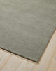 The corner of a grey green floor rug with a linear pattern in 100% wool with soft off white fringed edges. 