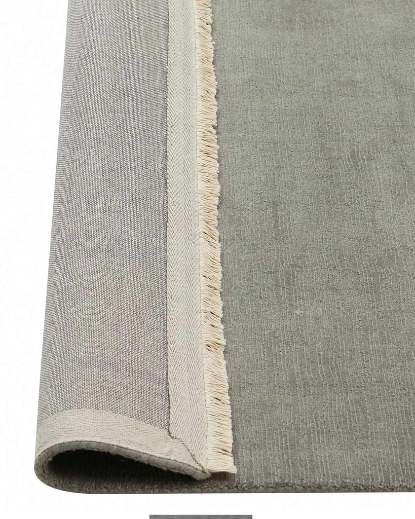A grey green 100% wool floor rug with cream fringe edges, the corner of which is flipped up to show the cotton backing. 