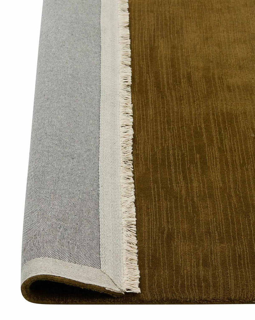 A detailed view of the linear weave of a 100% wool floor rug in deep yellow green brown tones, with a cream fringed edge. The corner is flipped up to show the sturdy cotton backing. 