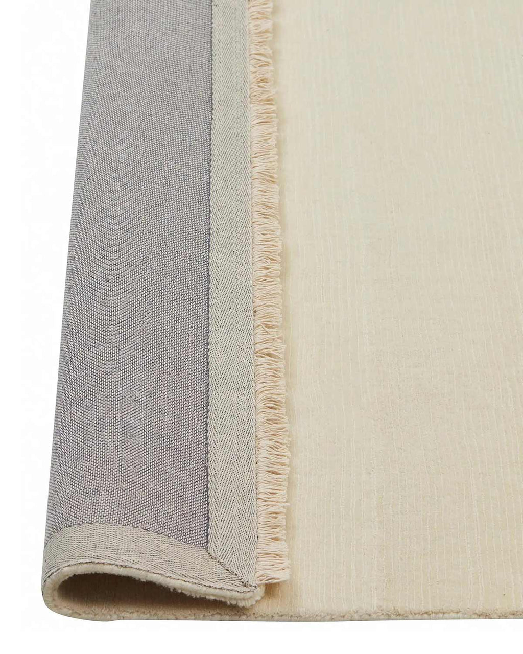 A view of the corner of a 100% wool cream toned floor rug with linear detailing and an off-white fringe, the edge of which is flipped up to show the sturdy cotton backing. 