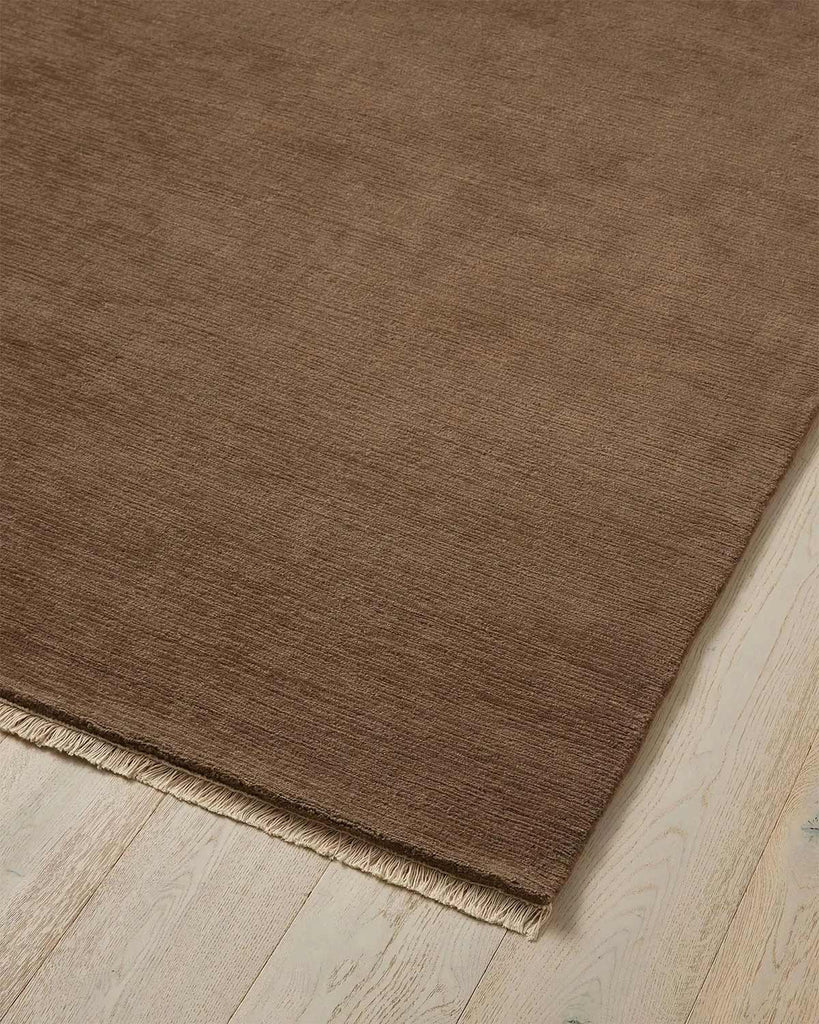 A corner image of a cool beige brown floor rug in 100% wool with slight linear texture in the weave and a cream fringed edge. 