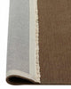 A corner of a deep taupe, cool brown floor rug with cream fringe at the edges in 100% wool with the corner flipped up to show the sturdy cotton backing. 