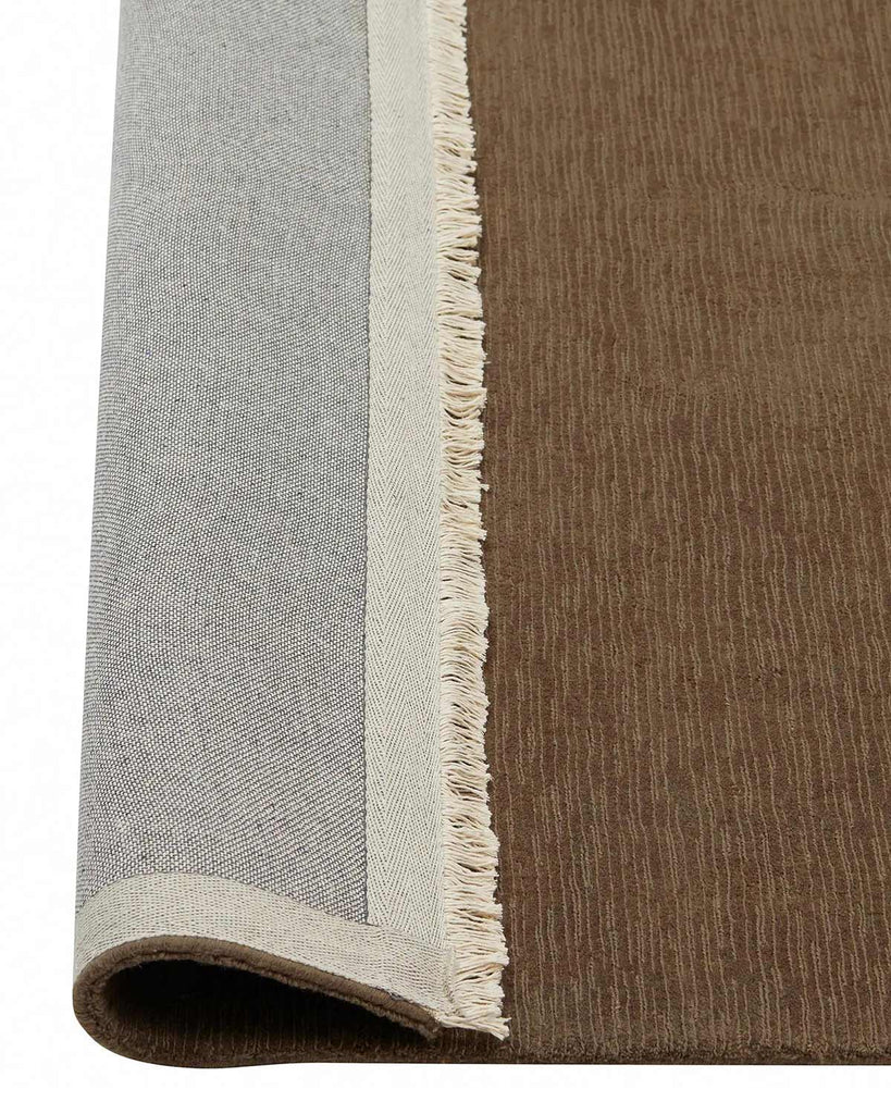 A corner of a deep taupe, cool brown floor rug with cream fringe at the edges in 100% wool with the corner flipped up to show the sturdy cotton backing. 