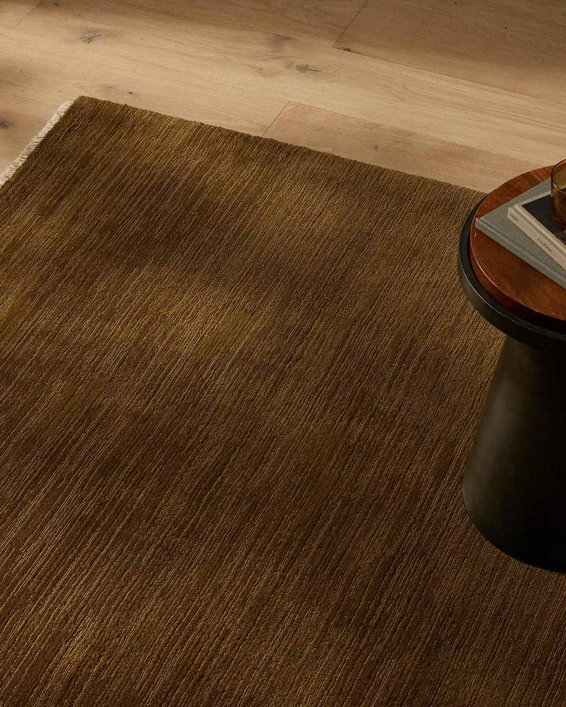 A deep green toned ochre floor rug with linear detailing (100% NZ wool) and cream toned fringe edges, in a stylish modern living room.