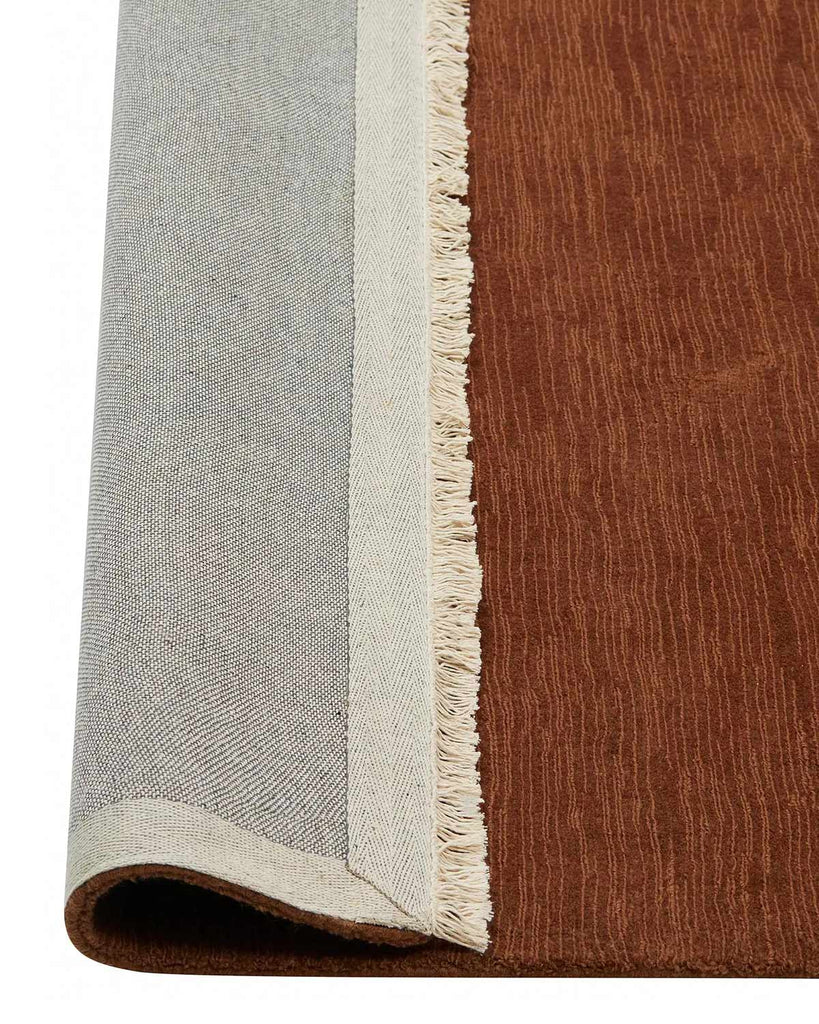 A red brown floor rug with cream toned fringed edges. The corner is flipped up to show the underside. 