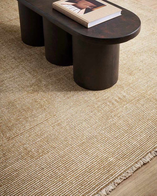 A coffee table on a warm toned beige, lightly distressed 100% NZ wool floor rug with a subtle fringed edge. 