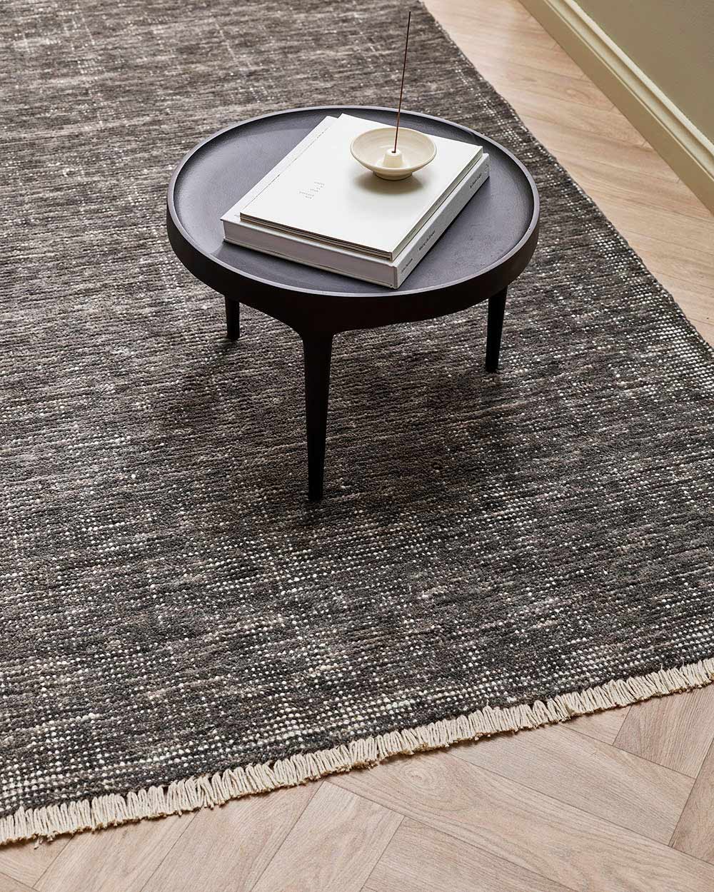 An edgy, slightly distressed 100% wool floor rug in a charcoal peat tone, with white detailing including a subtle fringed edge. 