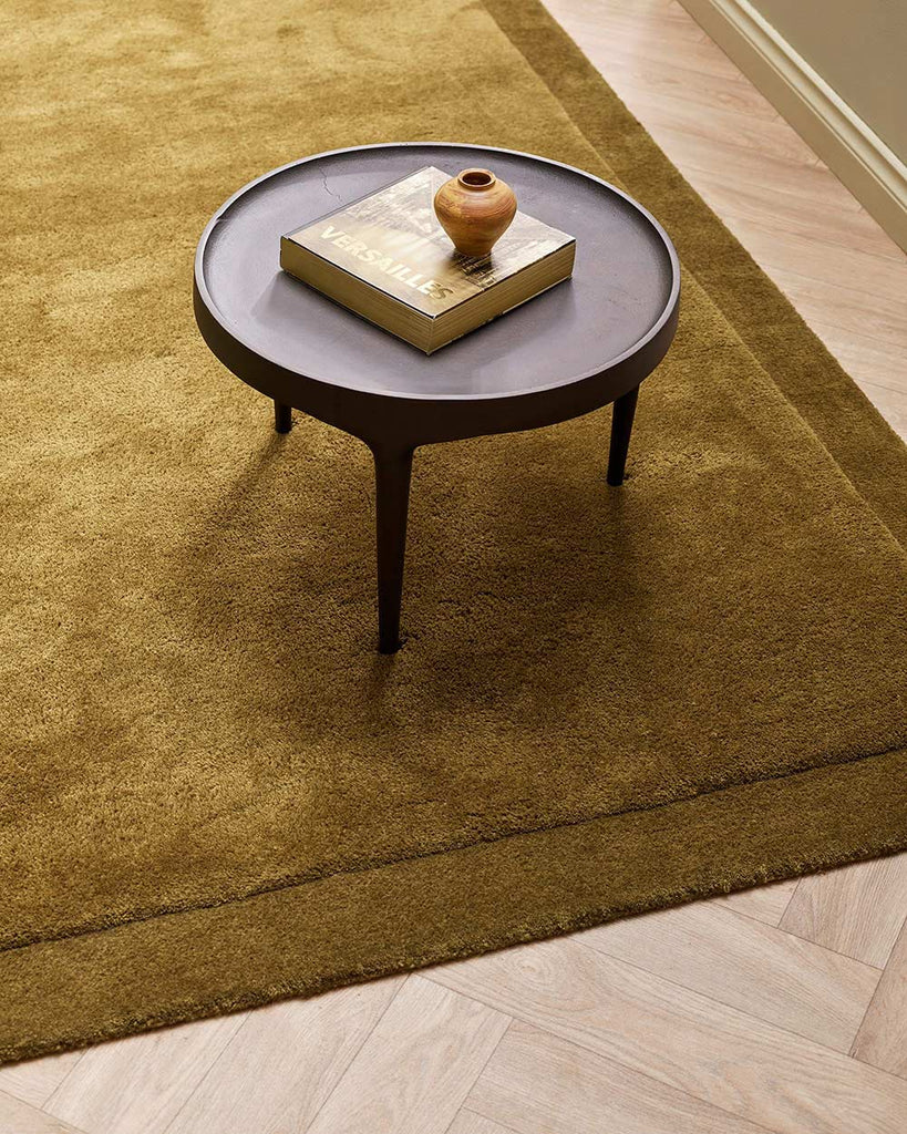 A coffee table on top of a luxurious warm green toned floor rug in 100% NZ wool with cut pile frame detailing along the edges, by Baya. 